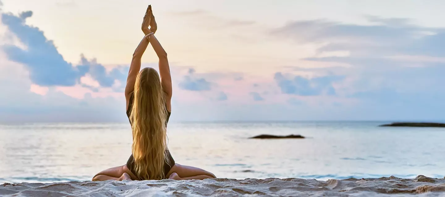 A young woman at the beach, doing yoga in a kneeling position with hands towards the sky and palms together, her back is turned to us with long hair flowing down her back